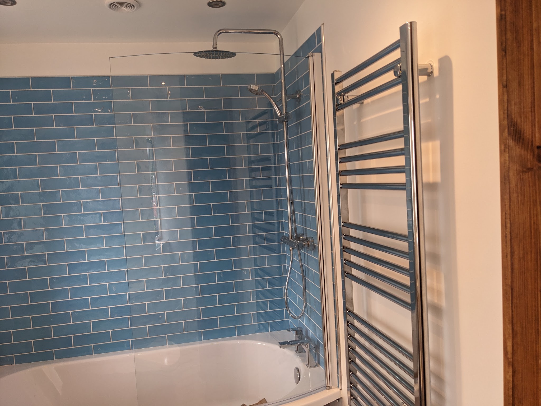 A bath with a curved glass screen against blue metro tiles, with a chrome towel rail beside it