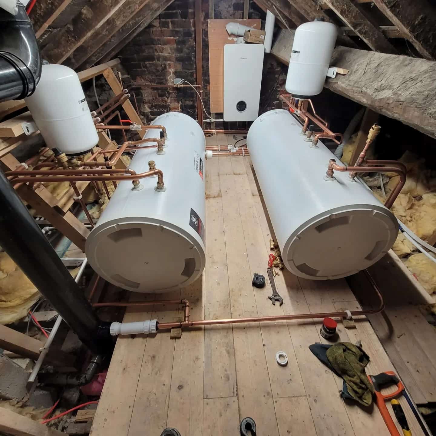 Two white hot water cylinders on a boarded loft floor, joined with copper pipework